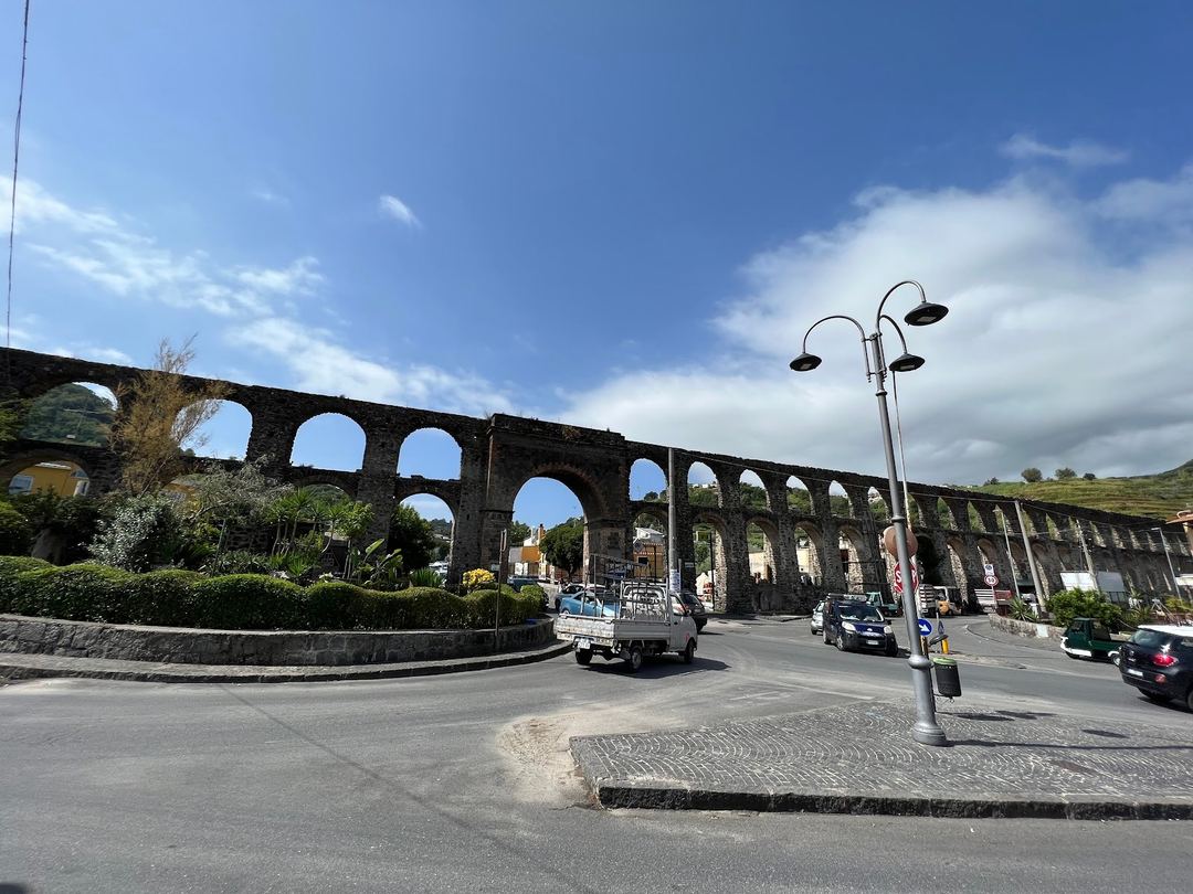 A historic Roman aqueduct with multiple arches spans across a street in what appears to be a Mediterranean town, with modern traffic and a street lamp in the foreground.