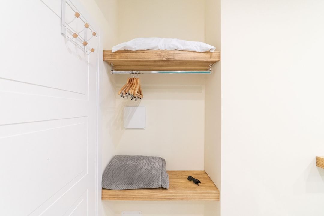 A compact built-in alcove bed with wooden shelving, white pillow, gray blanket, and wooden hangers for guest clothing storage.