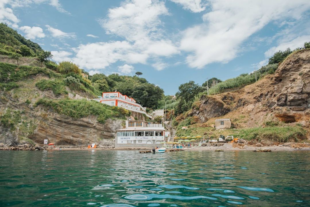Mandra B&B's waterfront location nestled between dramatic rocky cliffs with its distinctive orange and white building visible on the hillside above the beach.