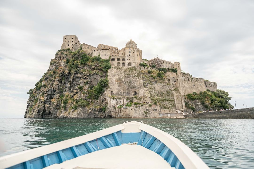 A scenic boat view of Aragonese Castle, a historic fortress perched on a volcanic rocky island in the Tyrrhenian Sea near Ischia, Italy.