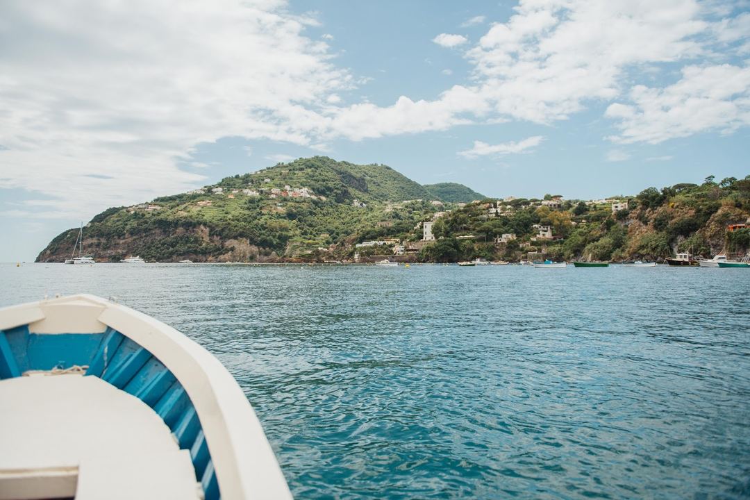 A scenic waterfront view of a lush hillside village taken from a boat, featuring turquoise Mediterranean waters and residential buildings nestled among green vegetation.