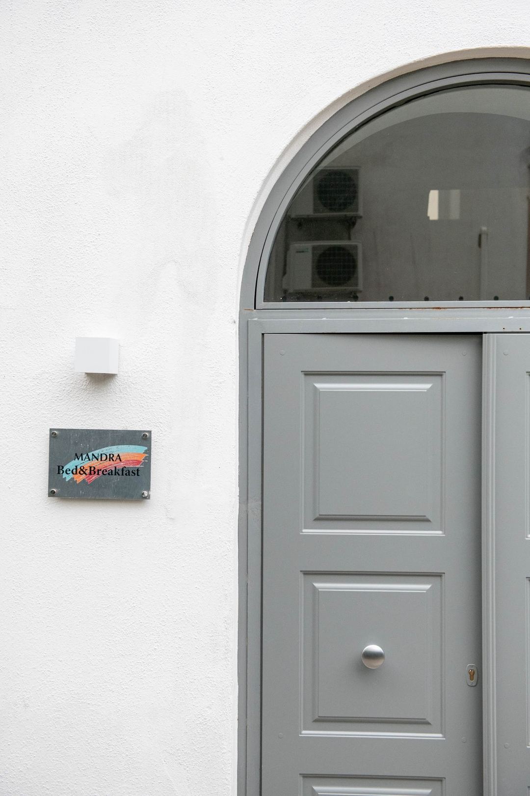 The entrance door of Mandra Bed & Breakfast featuring a gray arched doorway with a nameplate on the white-washed exterior wall.